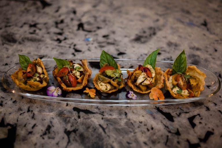 Five edible flower-garnished appetizers with basil leaves served on a long glass plate, set on a speckled stone surface.