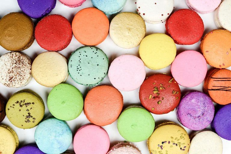 An assortment of brightly colored macarons with various flavors and sprinkled toppings, arranged in a grid pattern.