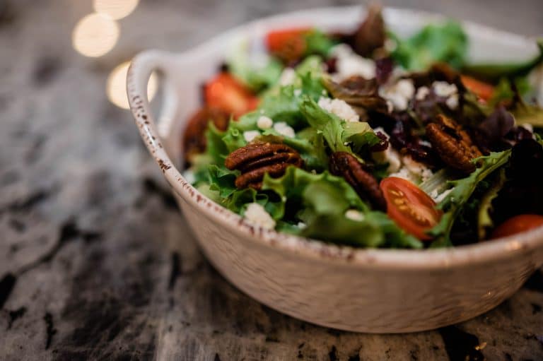 A white ceramic bowl filled with a salad, including mixed greens, cherry tomatoes, pecans, and crumbled cheese, placed on a marble countertop.