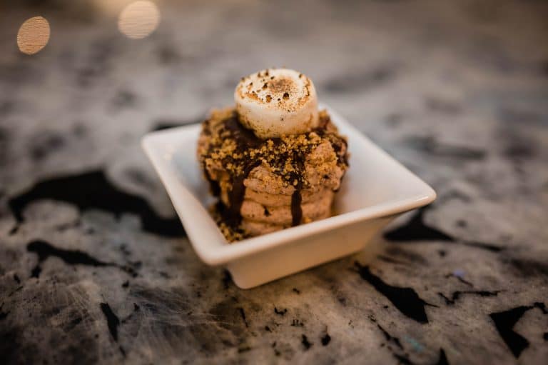 A dessert topped with a toasted marshmallow, chocolate drizzle, and crushed graham crackers, served in a small square dish on a marble surface.