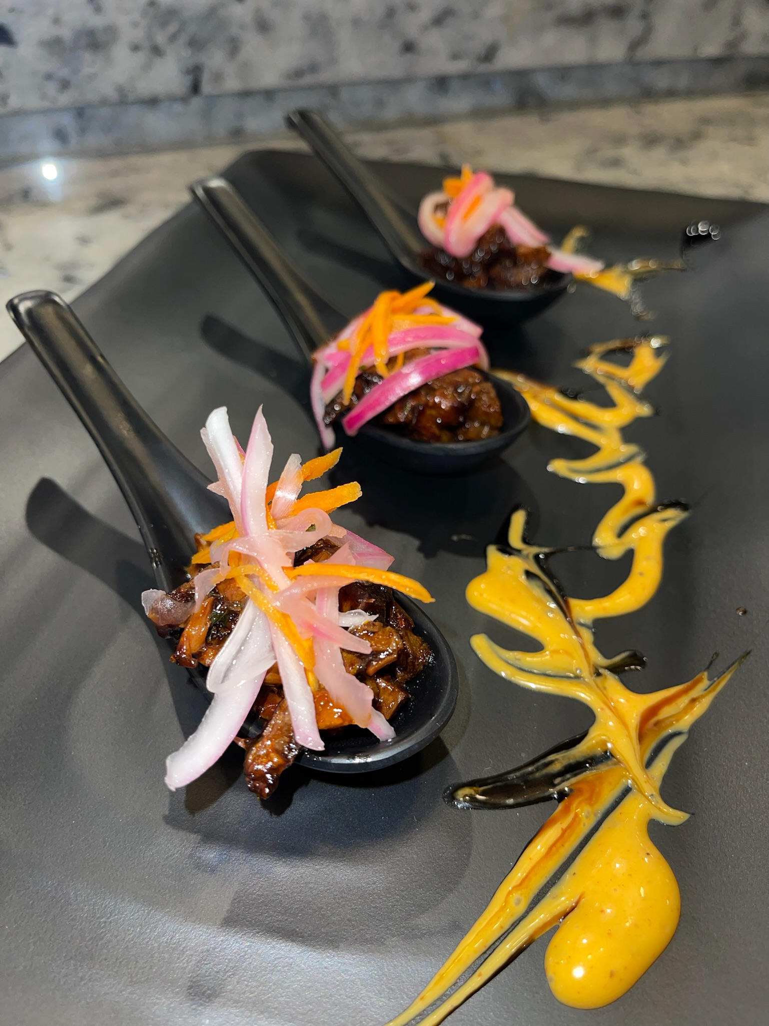Three black spoons of bite-sized appetizers garnished with pickled onions and carrots are arranged on a black plate with artistic drizzles of yellow and black sauce.