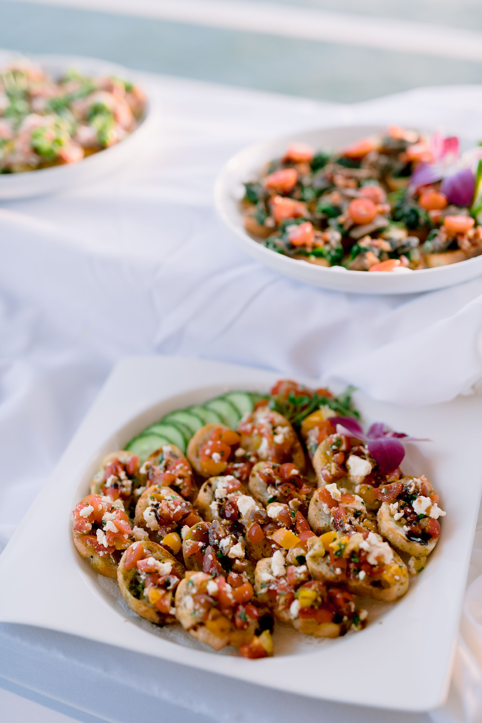 A plate of bruschetta topped with tomatoes, cheese, and herbs sits on a white tablecloth. Two additional bowls with mixed salads are in the background.