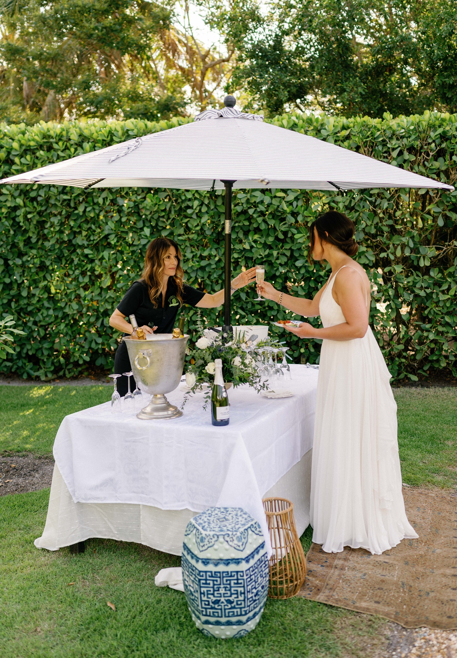 Two women clink glasses under a white umbrella at an outdoor event. A table with a white tablecloth, flowers, and a champagne bucket is set before them.