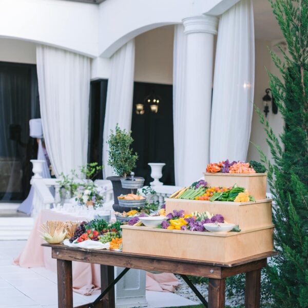 An outdoor event setup features a wooden table with a tiered display of assorted vegetables and fruits. In the background, there are white columns and draped curtains.