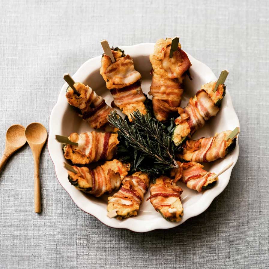 A white plate with nine pieces of bacon-wrapped chicken skewers arranged in a circle, garnished with rosemary. Two wooden spoons are placed to the left of the plate on a light gray tablecloth.