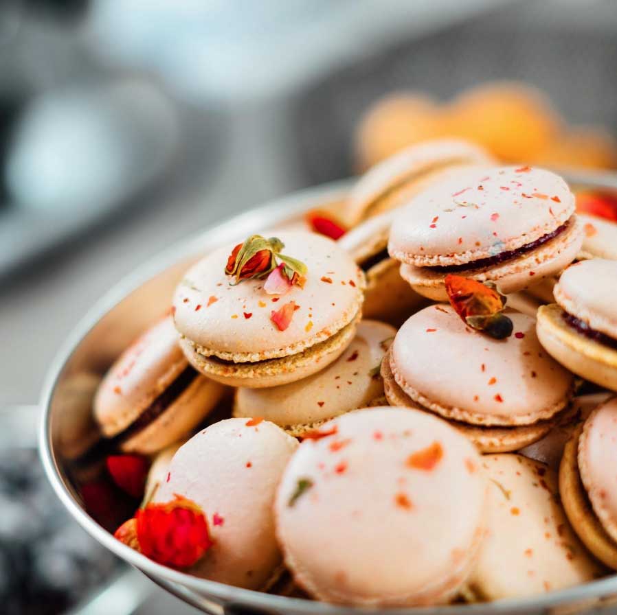 A bowl filled with white macarons, sprinkled with red and green toppings, and decorated with small dried rose petals.