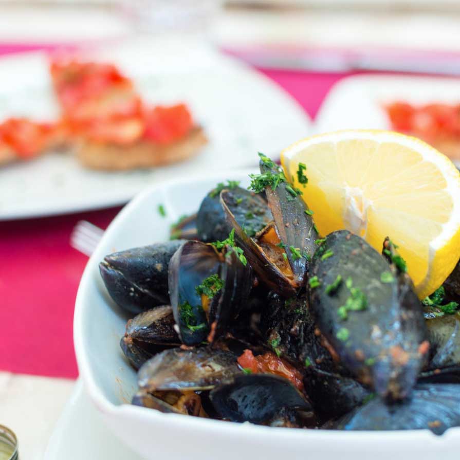 A bowl of cooked mussels garnished with chopped herbs and a lemon wedge, with bruschetta in the background.