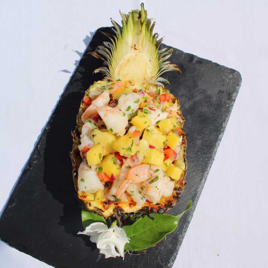 A halved pineapple filled with a shrimp and mango salad, garnished with herbs, and placed on a black slate.