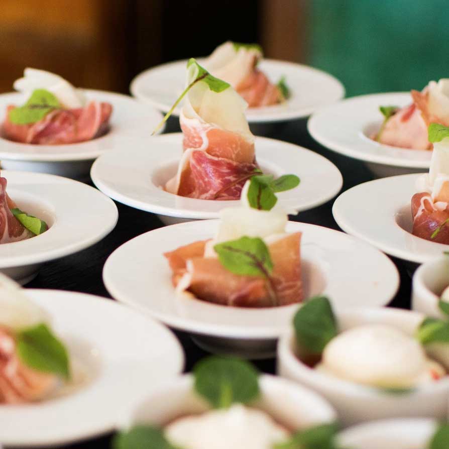 Rows of small white plates each holding a delicate arrangement of cured meat slices and greens, ready for serving.