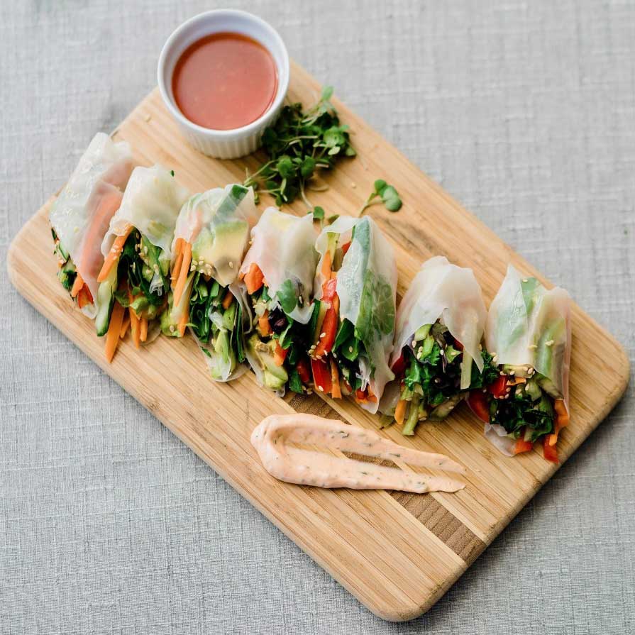 A wooden cutting board holding neatly arranged vegetable spring rolls, a small bowl of dipping sauce, and a dollop of a creamy condiment on a gray cloth background.