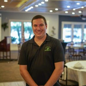 A man in a black polo shirt with a green logo stands smiling in a well-lit restaurant dining area.