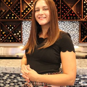 A person with long hair and a black shirt stands in front of a marble counter, smiling, with a background of wine bottles displayed in wooden racks.