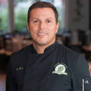 A chef in a black uniform stands inside a restaurant, looking at the camera with a slight smile. The background shows dining tables and chairs.