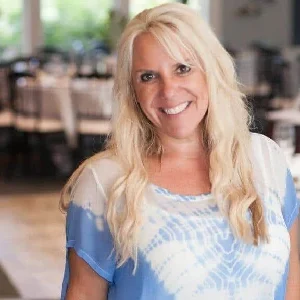 A woman with long blonde hair wearing a blue and white blouse stands in a restaurant and smiles at the camera.