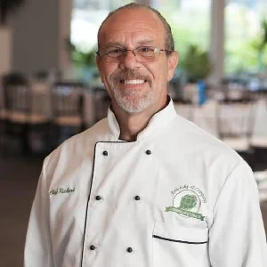 A smiling man in a white chef's coat stands in a dining area with neatly arranged tables in the background.